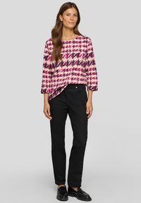 Frau mit langem braunem Haar, die eine Bluse mit rosa und lila Hahnentrittmuster, schwarze Hose und schwarze Loafers trägt und vor einem grauen Hintergrund steht.