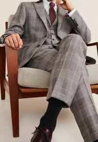 Completo a quadri grigio con gilet a tre bottoni e rever a punta, seduto su una sedia di legno. Scarpe in pelle marrone, calzini neri e dettagli sottili.