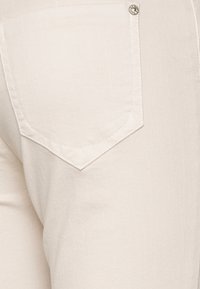 Jeans de mezclilla color beige claro con un bolsillo trasero de diseño puntiagudo y un acento de botón metálico. Textura suave y forma ajustada.