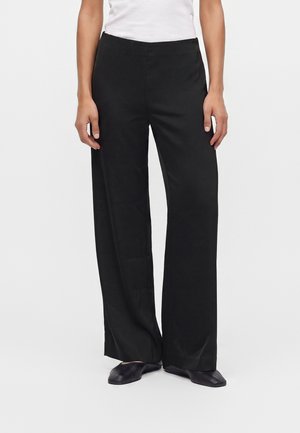 Personne portant un pantalon noir à jambes larges, un haut blanc et des chaussures plates noires, debout avec les bras détendus le long du corps devant un fond uni.