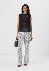 Top senza maniche in pizzo nero con bordi ondulati, abbinato a pantaloni bianchi con motivi che presentano linee astratte nere. La modella tiene una piccola borsa nera.