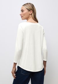 Weißes Langarmhemd mit weicher Textur, Rundhalsausschnitt und abgerundetem Saum, kombiniert mit dunklen Jeans. Der Fokus liegt auf der Schlichtheit und dem Design des Hemdes.