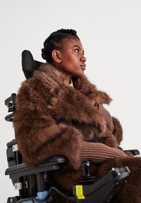 Γυναίκα με καφέ γούνινο παλτό, καθισμένη σε ηλεκτρικό αναπηρικό καρότσι, κοιτάζει σκεπτικά προς τα δεξιά σε ένα απλό φόντο.