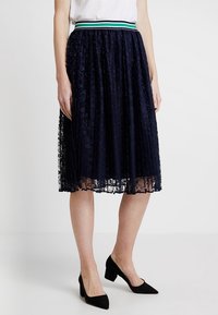 Granatowa koronkowa spódnica plisowana z szerokim pasem w zielono-białe paski, o zwiewnym kroju i delikatnej strukturze. Czarne szpilki dopełniają stylizację.