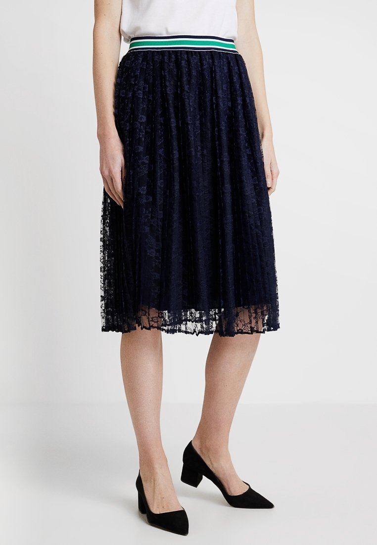Granatowa koronkowa spódnica plisowana z szerokim pasem w zielono-białe paski, o zwiewnym kroju i delikatnej strukturze. Czarne szpilki dopełniają stylizację.