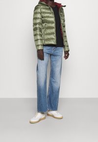 Kurtka puchowa w oliwkowej zieleni z kapturem i burgundowym wnętrzem. Połączona z jasnoniebieskimi spodniami typu wide-leg i białymi sneakerami.