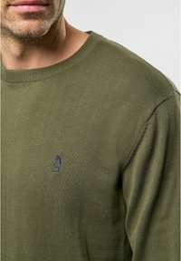 Pulover tricotat de culoare verde măsliniu cu guler rotund, prezentând un mic logo albastru pe piept și un model texturat al țesăturii.