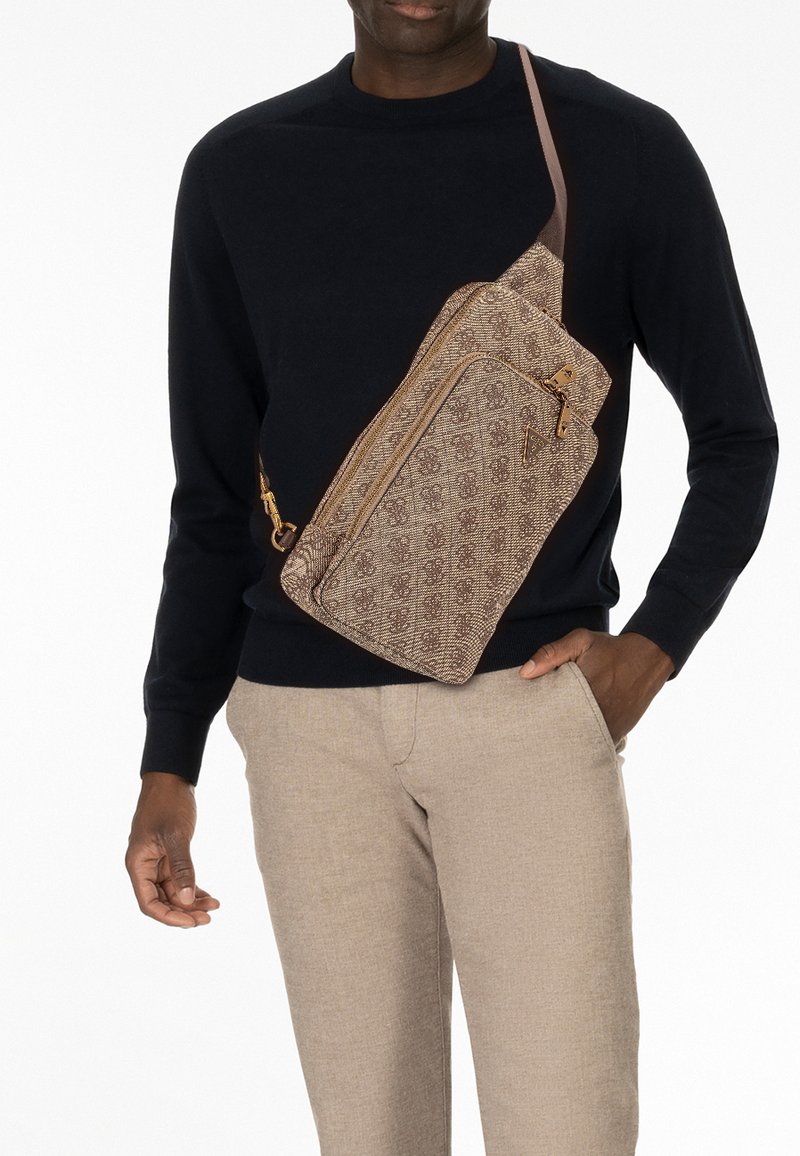 Borsa a tracolla marrone con motivo a fantasia, dalla forma curva, con chiusura a zip e tracolla. Indossata sopra un maglione scuro e pantaloni beige.