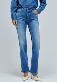 Vaqueros de denim azul con corte recto, diseño de cinco bolsillos y cierre de botón. Llevados con tacones bajos y puntiagudos de color claro.