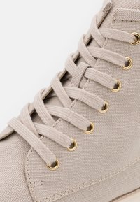 Gros plan d’une chaussure montante en tissu beige avec des lacets assortis passés dans des œillets métalliques dorés sur fond blanc.