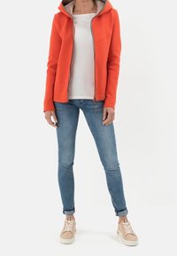 Frau trägt einen orangefarbenen Reißverschluss-Hoodie, ein weißes T-Shirt, enge blaue Jeans und beige Sneaker und steht vor einem weißen Hintergrund.