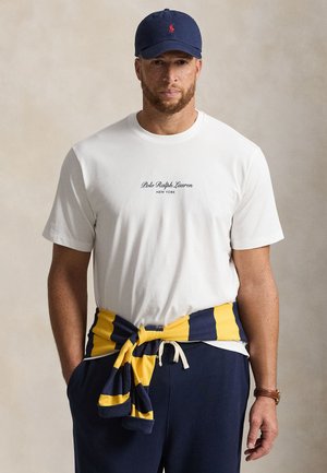 Mężczyzna ubrany w białą koszulkę Polo Ralph Lauren, granatową czapkę, granatowe spodnie oraz żółto-granatowy sweter w paski związany wokół talii.