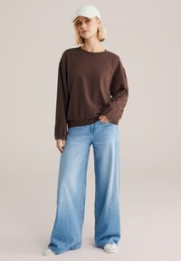 Ruda džemperis su ilgomis rankovėmis ir apvalia iškirpte, derinamas su šviesiai mėlynomis platėjusiomis džinsinėmis kelnėmis. Aprangą papildo šviesi kepurė ir kasdieniai sportbačiai.