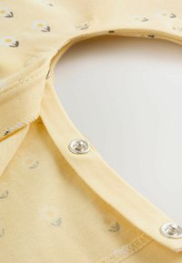 Gul bomullsbody med litet blommönster. Knäppning vid kragen. Mjuk textur med slät yta och rundad halsringning.