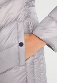 Lys grå dunjakke med kviltet design, en sidelomme med knaplukning og en glat, blank stofstruktur.