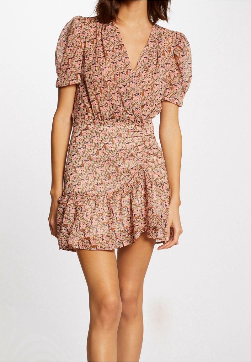 Femme portant une robe courte rose à motifs avec des manches bouffantes et un ourlet à volants, debout devant un fond blanc.