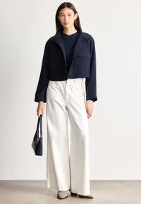 Femeie în picioare, purtând o jachetă scurtă bleumarin, pulover negru, pantaloni largi albi, pantofi bej ascuțiți, ținând o geantă mică bleumarin.