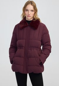 Donna bionda che indossa un giubbotto piumino viola scuro con colletto in pelliccia e le mani in tasca, su uno sfondo chiaro uniforme.