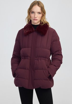 Blonde vrouw draagt een donkerpaarse pufferjack met een pluche kraag en heeft haar handen in haar zakken tegen een effen lichte achtergrond.