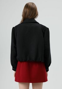 Zwarte cropped jas met opgeblazen mouwen en een puntige kraag; voorzien van een elastische zoom. Gecombineerd met een rode geplooide rok.