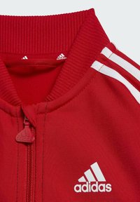 Chaqueta deportiva roja con cuello acanalado, cierre de cremallera y acentos en blanco con tres rayas en las mangas. Presenta un logo de Adidas en el pecho.