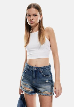 Un crop top blanc à côtes associé à un short en denim taille haute déstructuré avec des bords effilochés et un dégradé bleu clair.