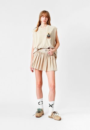 Top sin mangas de color beige con diseño de rayas y parche decorativo, combinado con una falda mini plizada de color beige. Se lleva con zapatillas beige y calcetines con patrones.