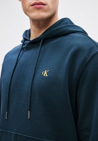 Felpa blu navy realizzata in materiale morbido, con logo cK ricamato in oro, cordino regolabile e tasca a marsupio sul davanti.