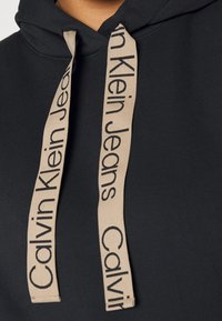 Černá mikina s kapucí, která má dvě tónované béžové tkaničky a potisk s textem "Calvin Klein Jeans." Látka vypadá hladce a má uvolněný střih.
