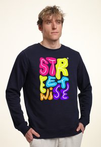 Námorně modrý svetr s kulatým výstřihem, na kterém je barevný text ve stylu bubliny "STREETWISE" v růžové, žluté, modré, oranžové a fialové barvě.