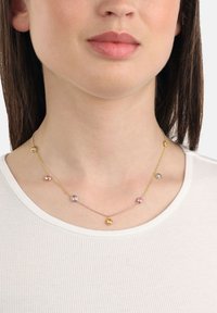 Donna che indossa una delicata collana di gemme multicolori con catene d'oro e d'argento intorno al collo nudo, visibile una maglietta bianca a costine.
