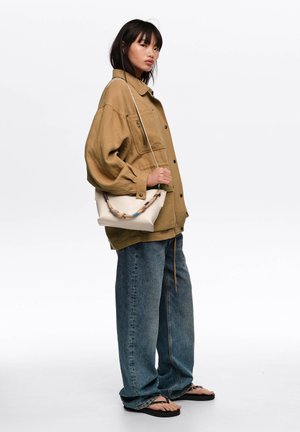 Mujer con chaqueta marrón clara holgada, vaqueros azules sueltos, chanclas negras y bolso blanco bandolera con asa decorativa, de pie contra fondo blanco.