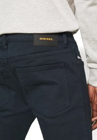 Primer plano de unos vaqueros Diesel de color azul marino combinados con un sudadera gris clara, mostrando el bolsillo trasero y la etiqueta de la marca en la cinturilla.