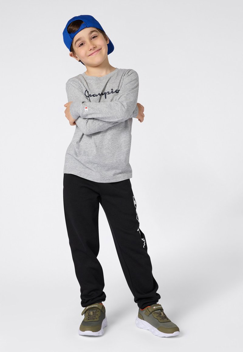 Uśmiechnięty chłopiec w niebieskiej czapce założonej tyłem, w szarym longsleeve'ie, czarnych dresowych spodniach i zielonych sneakersach, stojący z założonymi na siebie rękami.