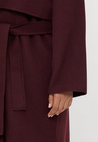 Maroon wrap coat with a belt, long sleeves, and soft, textured fabric. Features side seams and a clean finish at the edges.