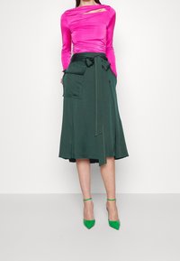 Žena ve světlé růžové dlouhém rukávu s výřezem a tmavě zelené midi sukni s velkými kapsami, doplněná zelenými vysokými podpatky.