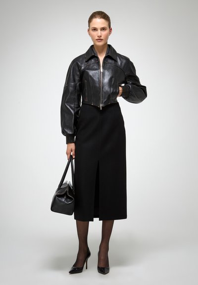 Chaqueta de cuero negra corta con un cuello alto y cierre de cremallera, combinada con una falda negra hasta la rodilla con una abertura en la parte delantera. Tacones negros y bolso.