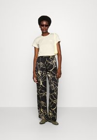 Camiseta de algodón beige con un pequeño emblema, combinada con pantalones de pierna ancha negros con un patrón floral amarillo. Zapatos de lona verde oliva.