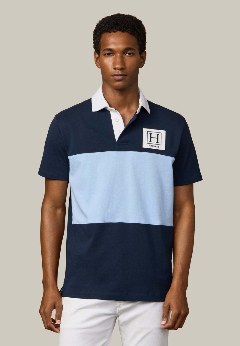 Jeune homme portant un polo rayé bleu marine et bleu clair avec col blanc et écusson, associé à un pantalon blanc sur un fond uni.