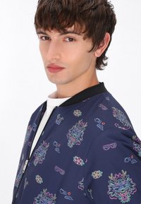 Giacca bomber della marina con vivace stampa grafica multicolore di fiori e occhiali da sole; colletto a coste nero e dettaglio con chiusura a zip.