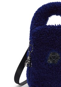 Bolso de mano azul de peluche con asa redondeada, correa negra, llavero de pequeño robot metálico y logo circular con una estrella y el nombre de la marca.