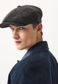 Siva kapa pepita uzorka napravljena od vune, s blago zaobljenom krunom i tamnom vrpcom, koju nosi model u tamnoplavom sakou, na bijeloj pozadini.