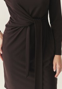 Vestido marrón oscuro de tela suave, con un detalle de nudo en la cintura y mangas largas, con un corsé ajustado y una falda fluida.