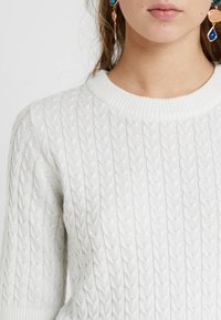 Valkoinen neulottu villapaita, jossa on teksturoitu punoskuvio ja resorireunuksinen pyöreä kaulus. Lähikuva, joka näyttää pehmeän materiaalin ja yksityiskohtaiset saumat.