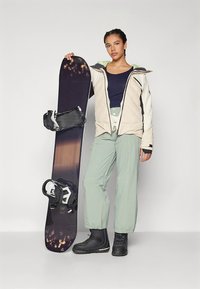 Snowboard mit einem dunkelvioletten Farbverlauf und schwarzen Bindungen; Modell trägt eine beige Jacke, mintgrüne Hose und schwarze Stiefel, steht vor einem neutralen Hintergrund.