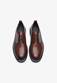 Chaussures en cuir marron avec une finition lisse, des lacets élastiques et une semelle en caoutchouc noir. Présente un design minimaliste et une forme épurée.