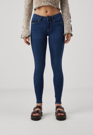 Personne portant un jean skinny bleu foncé taille haute, un pull court multicolore et des sandales noires à boucles, debout devant un fond uni.