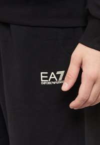Pantaloni della tuta neri realizzati in misto cotone, con un piccolo logo EA7 Emporio Armani bianco. Primo piano della tasca laterale e dei dettagli delle cuciture.