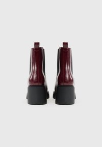 Botas de tornozelo em pele bordô com um salto preto robusto, painéis laterais e alças na parte de trás para facilitar o calçar. Textura suave e brilhante.