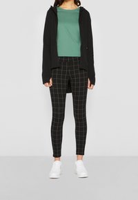 Černá mikina na zip s dlouhými rukávy, zelené tričko a černé kostkované legíny. Bílé tenisky dokončují tento outfit.